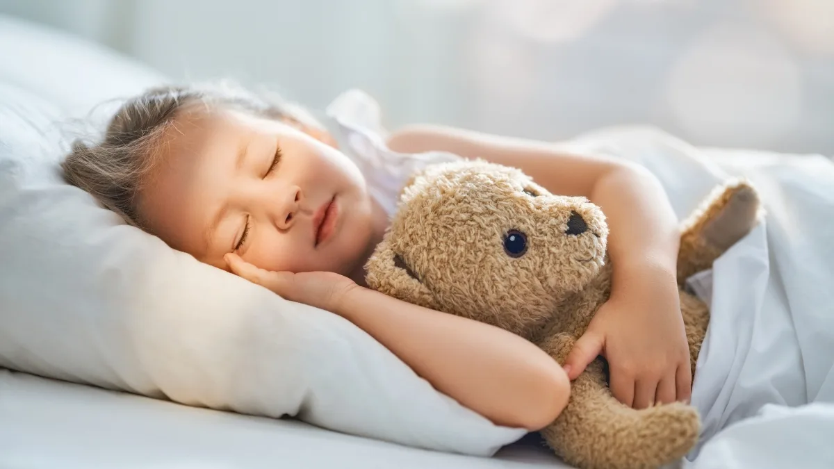 Enfant endormi sur un oreiller blanc, serrant contre lui un ours en peluche beige, dans une lumière douce